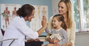 mom with daughter sitting in lap giving pediatrician high five; blog: 5 Reasons Well Child Visits are Important