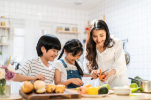 Lovely cute Asian family making food in kitchen at home. Portrait of smiling mother and children standing at cooking counter preparing ingredient for dinner meal. Happy family activity together; blog: Ways to Help Your Child with Weight Management