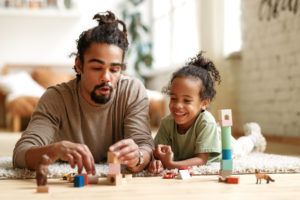 Happy african american family father and child son playing toys together at home, positive dad parent and little kid sitting on floor and stacking wooden pyramid, laughing while spending leisure time; The Importance of Play in Child Development